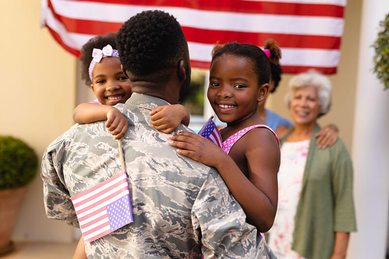 A soldier in uniform is seen from behind, holding his two young daughters who are smiling and holding small American flags. An American flag hangs in the background, and an older woman is seen smiling in the background, creating a warm, welcoming scene of a family reunion.