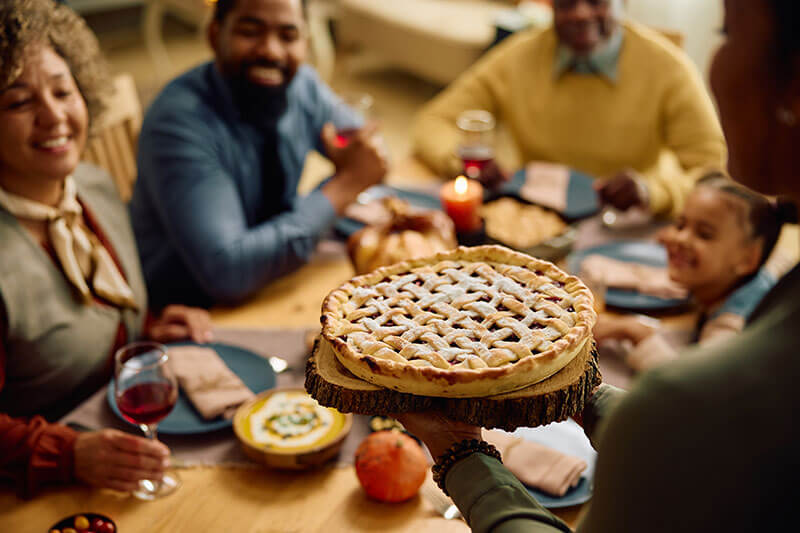 A close-up of a woman serving a homemade lattice-topped pie at a Thanksgiving family dinner. The family is gathered around a festively decorated table, smiling and enjoying the moment together.